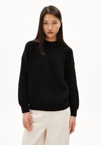 Schwarzer Strickpullover mit hohem Kragen, gerippten Bündchen und lockerer Passform. Der Stoff hat ein strukturiertes Muster. Kombiniert mit hellen Hosen.