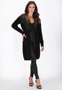 Frau trägt einen schwarzen langen Cardigan mit goldenen Pailletten auf den Schultern, ein schwarzes Top, schwarze Lederhosen, Creolen und schwarze High-Heels.