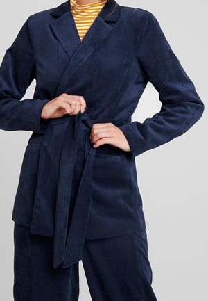 Personne attachant la ceinture d'un blazer en velours côtelé bleu marine porté sur un pantalon assorti et une chemise rayée jaune.