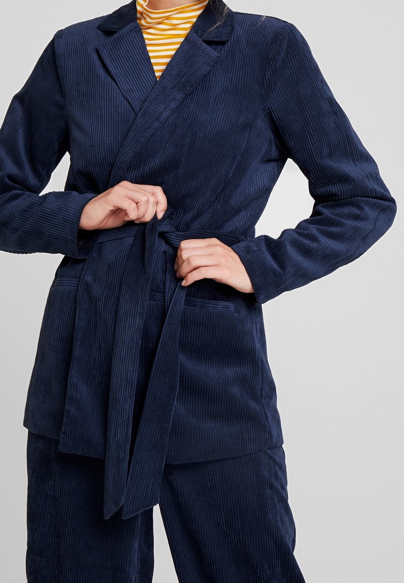 Personne attachant la ceinture d'un blazer en velours côtelé bleu marine porté sur un pantalon assorti et une chemise rayée jaune.
