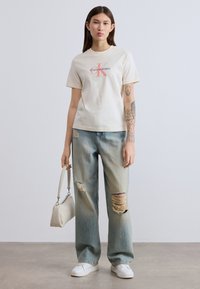 Μπεζ βαμβακερό T-shirt με τυπωμένο λογότυπο, σε συνδυασμό με ανοιχτό μπλε τζιν με σκισίματα, συνοδευόμενο από μικρή λευκή τσάντα και αθλητικά παπούτσια.