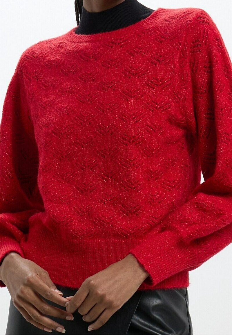 Personne portant un pull en maille rouge texturée avec un col haut noir, associé à un pantalon noir, les mains délicatement jointes à la taille.