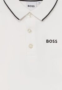 Biela polo košeľa z textúrovanej látky, s golierom s čiernym lemovaním, trojica gombíkov a čierne logo "BOSS" na hrudi.