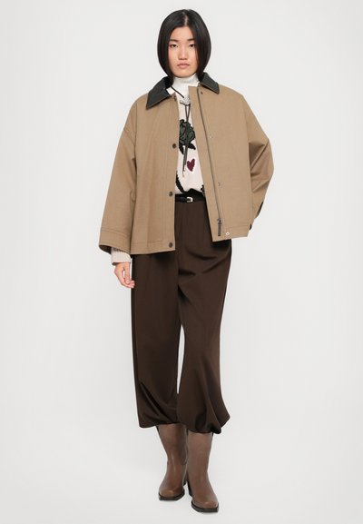 Veste oversized brune avec un col noir, pull blanc à motif floral, pantalon taille haute brun et bottines brunes.