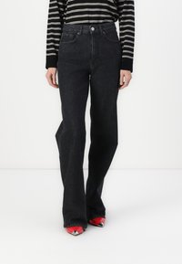 Black denim flared jeans with a high waist, five pockets, and a front button closure, paired with red pointed-toe shoes.