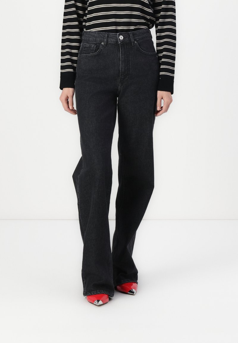Black denim flared jeans with a high waist, five pockets, and a front button closure, paired with red pointed-toe shoes.
