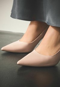 Alasti lakknahast kõrge kontsaga kingad terava varba ja madala pealiskattega, taustal tume tekstuuriga pind ja hall seelikusäär.