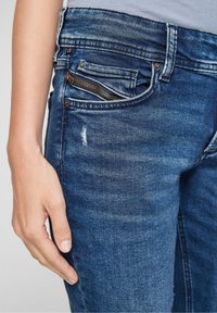 Vaqueros de mezclilla azul oscuro con un bolsillo frontal que cuenta con un zipper de bronce. La tela muestra un ligero desgaste y detalles desgastados.