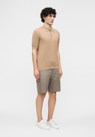 Junger Mann mit lockigem Haar, der ein beige kurzes Strick-Poloshirt, graue Shorts und weiße Turnschuhe trägt und vor einem schlichten weißen Hintergrund steht.
