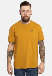 T-shirt di cotone giallo senape con maniche corte, colletto tondo, taschino sul lato sinistro e una piccola etichetta di marca. Indossato con jeans blu.