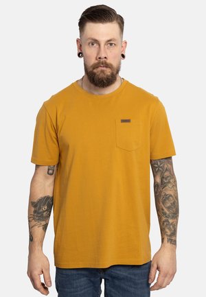 Mosterdgele katoenen T-shirt met korte mouwen, ronde hals, linkerk borstzak en een klein merklabel. Draagt bij blauwe spijkerbroek.