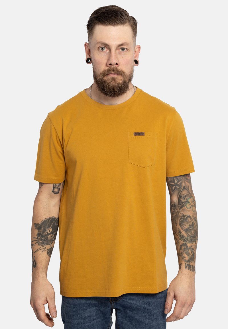 T-shirt di cotone giallo senape con maniche corte, colletto tondo, taschino sul lato sinistro e una piccola etichetta di marca. Indossato con jeans blu.