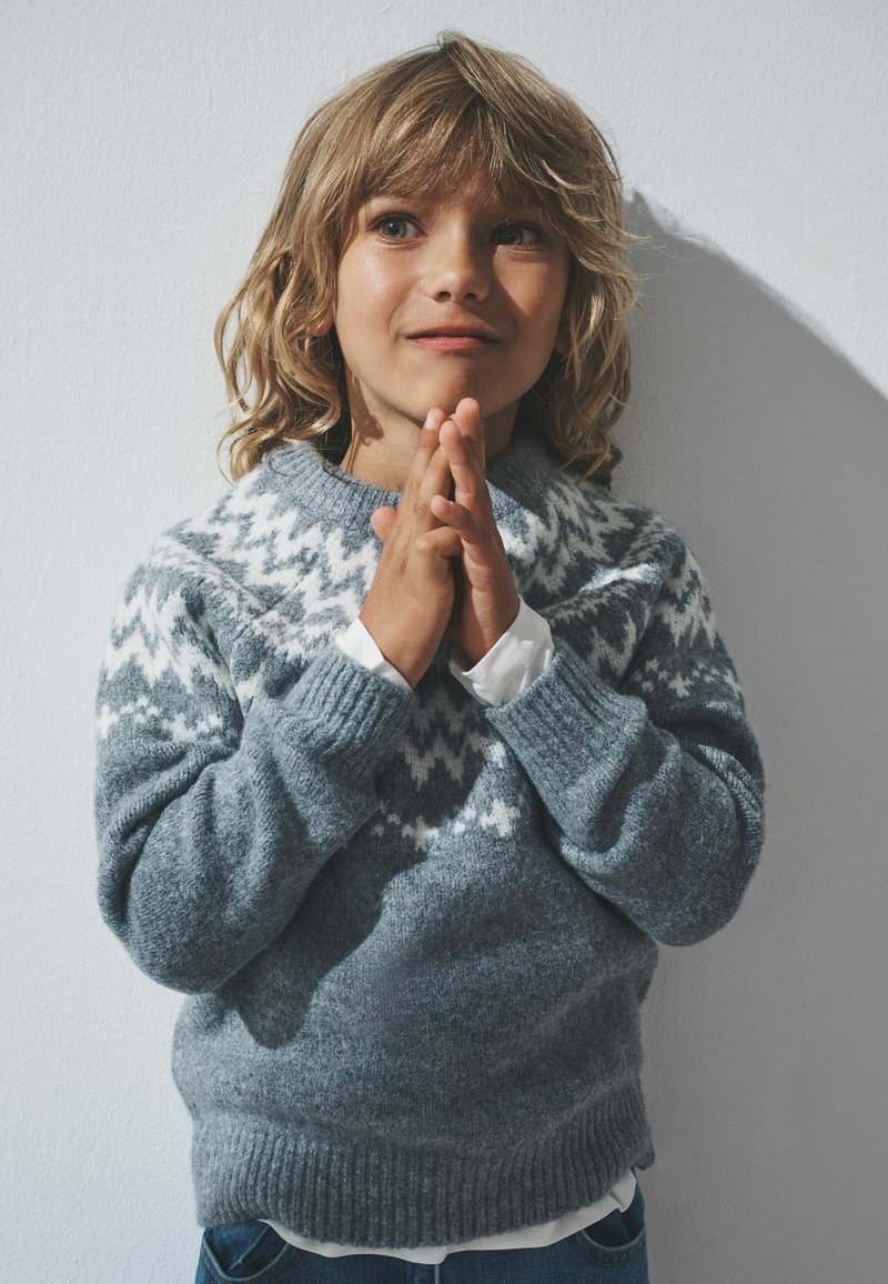 Maglione grigio a maglia con accenti bianchi a motivo, con collo rotondo e polsini a coste. Le mani del modello sono incrociate.
