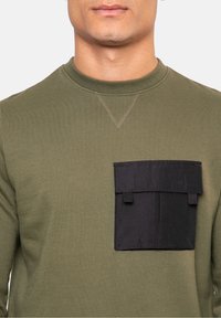 Olivově zelená mikina s texturovanou černou kapsou. Žebrovaný výstřih a viditelné šití, přiléhavý střih s dlouhými rukávy.
