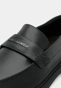 Μαύρο δερμάτινο μοκασίνι με στρογγυλερή μύτη, λεπτομέρειες με λουράκι και μεταλλικό λογότυπο. Λείο υλικό με διακριτικές ραφές στα άκρα.