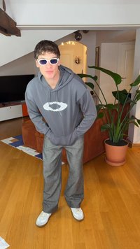 Grijze capuchontrui met een logo, brede grijze geruite broek, witte sneakers en oversized witte zonnebril. Binnenomgeving met planten.