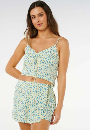 Joven mujer que lleva un top corto amarillo con estampado floral y una falda envolvente a juego, posando con una mano en la cadera sobre un fondo blanco.