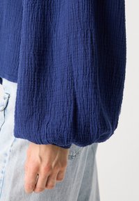 Navy blue textured top with long, gathered sleeves. The fabric features a crinkled pattern, paired with light blue denim bottoms.