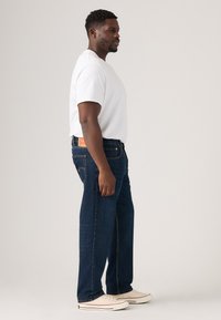 Mann, der seitlich steht und ein einfaches weißes T-Shirt, dunkelblaue Jeans und weiße Sneakers trägt, vor einem neutralen Hintergrund.