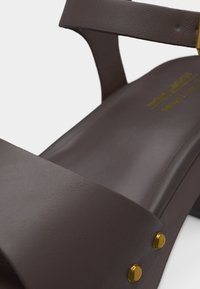 Κοντινό πλάνο σκούρου καφέ δερμάτινου σανδαλιού με χοντρό τακούνι, λουρί στον αστράγαλο και χρυσές λεπτομέρειες με καρφιά στο πλάι.