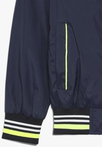 Námornícke bombardovací sako s elastickými manžetami, s neonově zeleným lemem a boční kapsou. Vyrobeno z hladkého, lehkého materiálu.