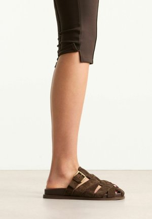 Partie inférieure de la jambe portant un pantalon court foncé et des sandales marron en daim à bout ouvert avec une bride à boucle, debout sur un sol clair.