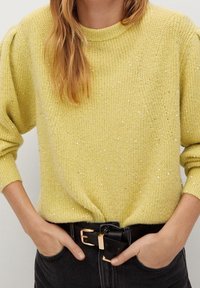 Gelber Strickpullover mit strukturiertem Finish und subtilen Glitzerakzenten, mit rundem Ausschnitt und langen Ärmeln, kombiniert mit einem schwarzen Gürtel.