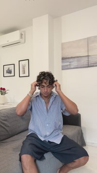 Šviesiai mėlynos ir baltos spalvų dryžuota trumpomis rankovėmis marškinėliai su mygtukais, derinami su tamsiai dryžuotomis šortais. Sėdi ant pilkos sofos, viduje.