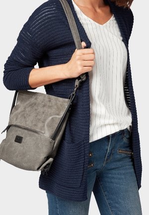 Kobieta ubrana w granatowy dzianinowy sweter rozpinany, białą bluzkę w paski, niebieskie dżinsy, niosąca szarą torbę na ramię na lewym ramieniu.