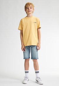 Gul bomulds T-shirt med trykt logo, parret med lysblå denim shorts. Hvide sneakers og stribede sokker fuldender outfittet.