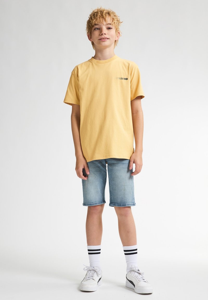 Gul bomulds T-shirt med trykt logo, parret med lysblå denim shorts. Hvide sneakers og stribede sokker fuldender outfittet.