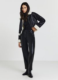 Femme portant une veste en shearling noir, un col roulé noir, un pantalon en cuir noir taille haute et des bottines noires, se tenant devant un fond blanc.
