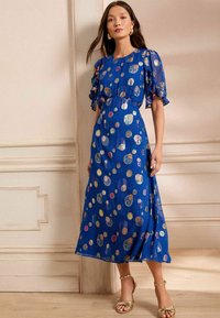 Modra obleka z nabranimi rokavi, okrašena z večbarvnimi krožnimi vzorci. Narejena iz lahkega materiala, seže do dolžine sredine tele. Zlate sandale.