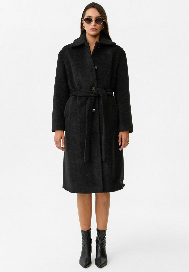 Zwart wollen blend jas met een kraag, lange mouwen, knopen aan de voorkant, een strikriem in de taille en zijzakken. Draag het met enkellaarsjes.