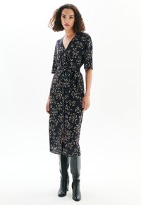 Femme portant une robe midi portefeuille noire à fleurs avec des manches aux coudes et des bottes à talons hautes noires jusqu'aux genoux, se tenant devant un fond blanc.