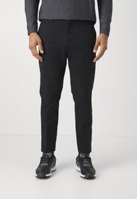 Pantalon noir slim en tissu lisse ; il dispose de passants de ceinture, de poches latérales et d'un design à l'avant plat, associé à des chaussures de sport noires.