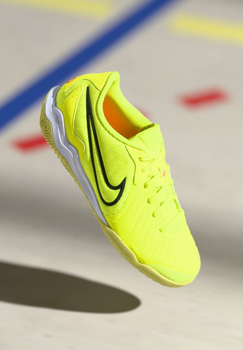 Scarpa da soccer indoor gialla brillante con il logo Nike nero e suola bianca, sospesa sopra un pavimento da palestra con linee colorate.