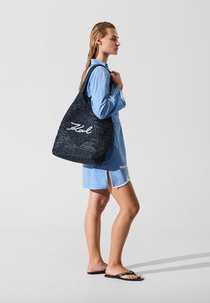 Sac cabas tissé bleu marine avec logo script blanc en grand format. Dispose de larges poignées et d'une ouverture supérieure. Le modèle porte une robe chemise bleu clair et des sandales.