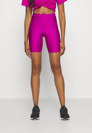 Vrouw draagt felroze, hooggetailleerde fietsshorts en een bijpassend geknoopt cropped topje, met zwarte sportschoenen, staand tegen een effen achtergrond.