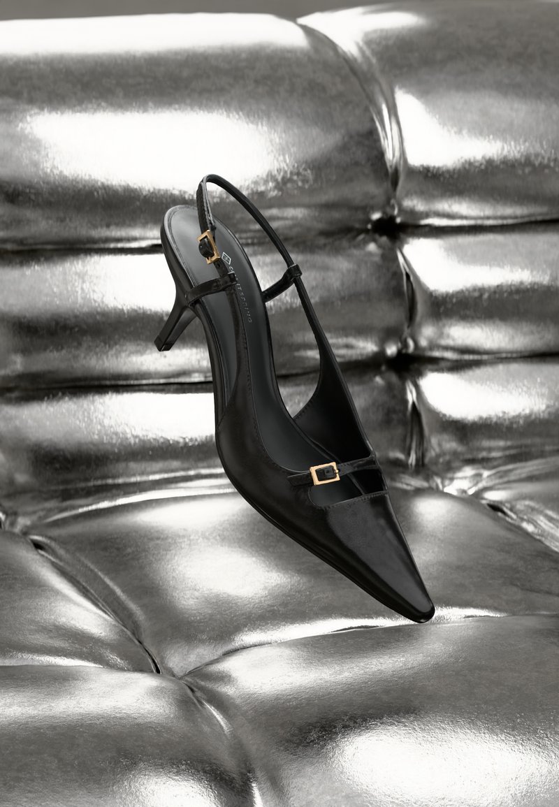Černé kožené slingback lodičky na nízkém jehlovém podpatku se zlatými sponami odpočívají na lesklém stříbrném polstrovaném povrchu.