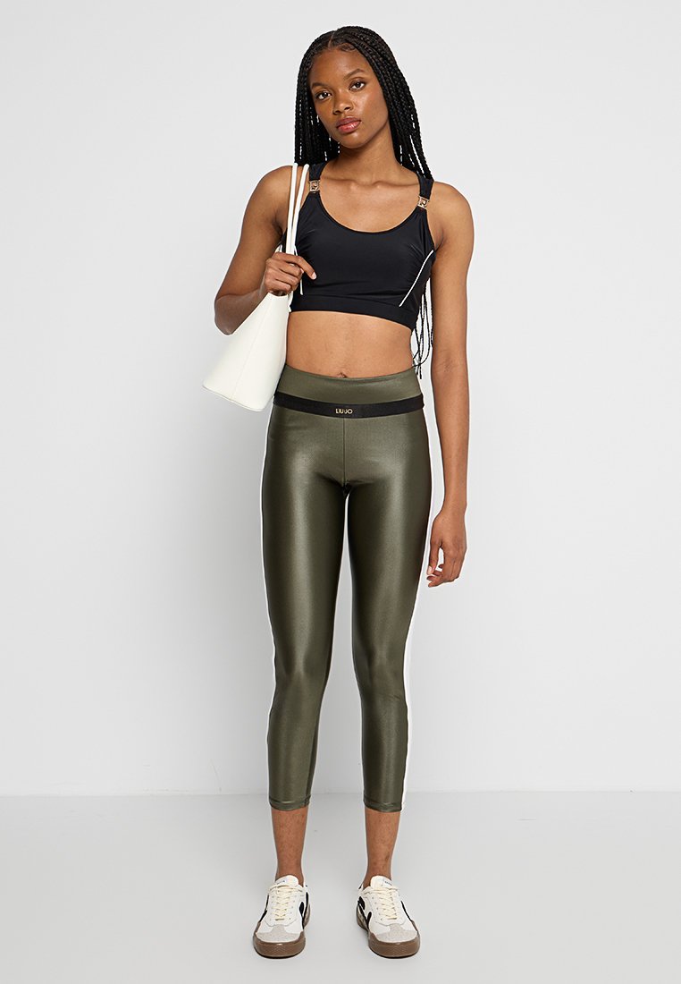 Czarny crop top na cienkich ramiączkach, zestawiony z oliwkowymi metalicznymi legginsami o wysokim stanie. Modelka trzyma białą torebkę.