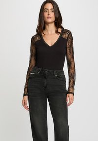 Kvinde iført sort V-hals top med gennemsigtig blonde lange ærmer og højtaljede mørkegrå jeans foran en ensfarvet baggrund.