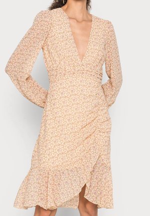 Kvinde iført en lyse ferskenfarvet blomsterkjole med lange ærmer, dyb V-udskæring, rynket nederdel og flæsekant mod en ensfarvet baggrund.