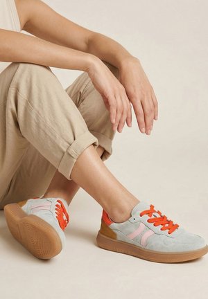 Osoba siedząca, ubrana w beżowe podwinięte spodnie i szare tenisówki z różowymi akcentami i jasnopomarańczowymi sznurowadłami na jednolitym tle.