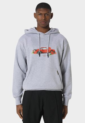 Junger Mann trägt einen hellgrauen Hoodie mit einem roten Oldtimer und roten Rosen auf der Brust, kombiniert mit schwarzen Hosen.