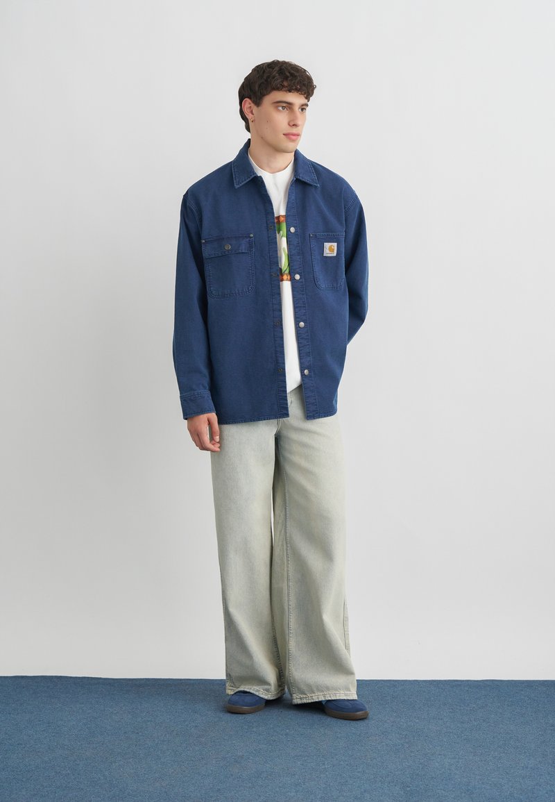 Junger Mann steht, trägt eine blaue Jacke mit Knöpfen, ein weißes T-Shirt mit Grafik, helle weite Jeans und blaue Schuhe, vor einer weißen Wand.