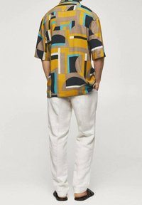 Camisa de manga corta con un patrón geométrico en beige, negro, amarillo y azul. Combinada con pantalones blancos y sandalias negras.