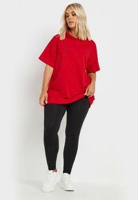 Röd oversized hoodie med en huva och korta ärmar, i kombination med svarta leggings och vita sneakers. Slät textur och avskalat design.