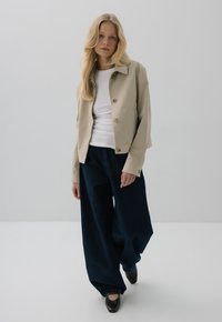 Beżowa krótka kurtka z guzikami, zestawiona z luźnymi ciemnoniebieskimi spodniami dżinsowymi i czarnymi butami. Gładkie tekstury i czysty design.