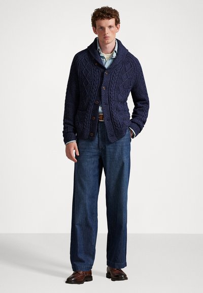 Hombre con un cárdigan de punto de cable azul marino, camisa de botones azul claro, jeans de pierna ancha y zapatos de cuero marrón, de pie contra un fondo blanco.